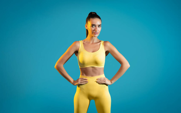 Fitness Workout. Fit Woman Wearing Sportswear Posing With Hands On Hips Looking At Camera Standing In Studio On Blue Background. Fitness And Gym, Sporty Lifestyle Concept - Photo, image