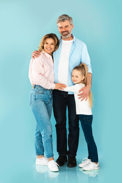 Happy Family Embracing Standing Together Smiling To Camera Posing Over Blue Studio Background. Middle Aged Parents And Little Daughter Hugging Expressing Love. Full Length, Vertical - Photo, image