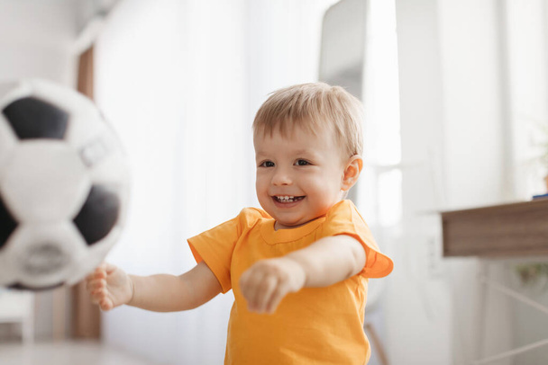 Радостный маленький мальчик, играющий с футбольным мячом, веселящийся и наслаждающийся игрой дома, интерьер гостиной. Очаровательный малыш бросает мяч и улыбается - Фото, изображение