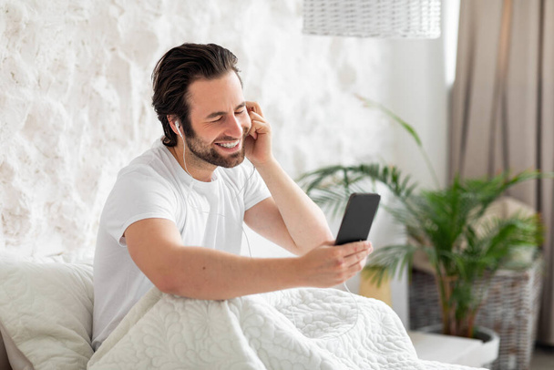 Fröhlicher bärtiger junger Kerl, der morgens nach dem Aufwachen Videoanrufe mit Freundin führt, glücklicher Millennial-Mann, der im Bett sitzt, Smartphone und Kopfhörer benutzt, auf den Gadget-Bildschirm schaut und lächelt - Foto, Bild