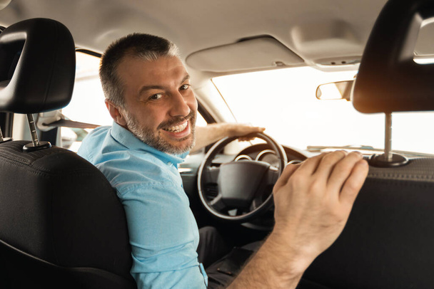 Cheerful man driving new automobile after purchase, sitting on drivers seat of car at dealership store shop. Happy guy looking back at passenger seat and smiling, buying modern auto at showroom - Fotoğraf, Görsel