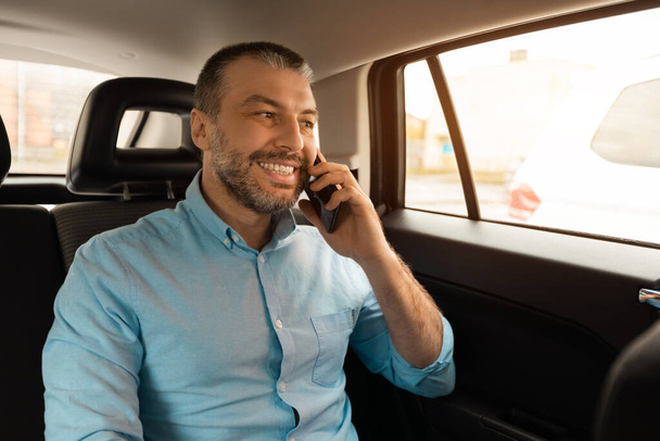 Portrait of happy businessman talking on mobile phone with clients, looking through window, going to business meeting by car, middle aged manager sitting on back seat, having conversation on cellphone - Photo, Image
