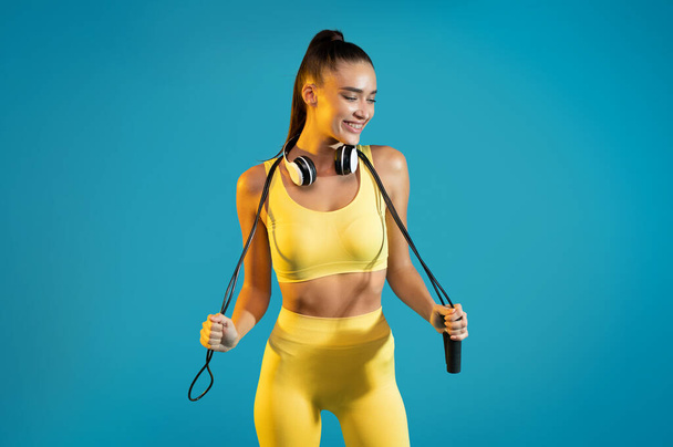 Glücklich Fit Frau in Sportbekleidung posiert mit Springseil beim Workout stehend im Studio Blue Studio Hintergrund, Blick zur Seite. Fitness und sportliches Lifestyle-Konzept - Foto, Bild