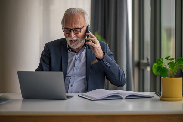 Anxious businessman talking on mobile phone while working on laptop. Worried senior male professional is wearing formals. He is sitting at desk in corporate office. - Фото, изображение