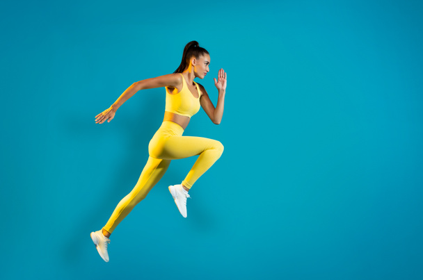 Side View Of Sportswoman Jumping In Mid Air Posing Looking Aside Exercising Over Blue Studio Background. Fitness Lady Running In Air. Sport Motivation. Full Length Shot - 写真・画像