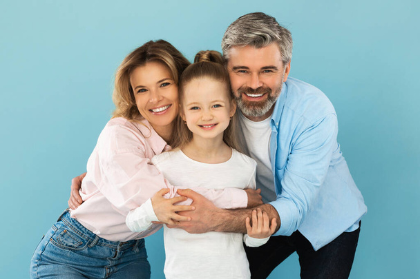 Felicidad familiar. Padres de la Edad Media felices abrazando a su pequeña hija posando de pie sobre el fondo del estudio azul, sonriendo a la cámara. Alegría del concepto de paternidad - Foto, Imagen