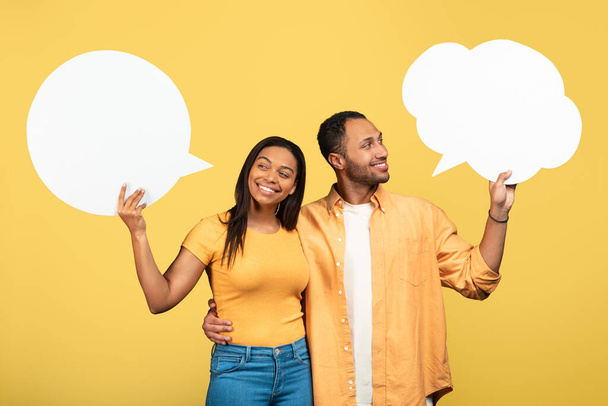 Feliz joven pareja negra sosteniendo burbujas de habla vacías en el fondo amarillo del estudio, maqueta. Guapo millennial afroamericano chico y su novia posando con palabras en blanco nubes - Foto, imagen