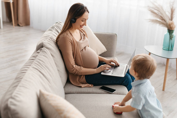 ノートパソコンを使用してヘッドセットで幸せな妊婦、勉強やオンラインで動作しながら、小さな息子が近くに再生します。リモートジョブと産休を楽しむ正の期待の母親 - 写真・画像