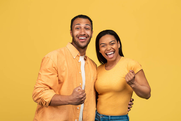 Excited young black man and woman gesturing YES, celebrating success on yellow studio background. Happy African American couple feeling overjoyed about huge sale or special offer - Фото, изображение