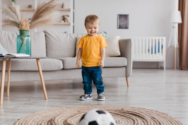 Πορτρέτο του αξιολάτρευτο παιδί αγόρι ποδοσφαιριστής παίζει μπάλα ποδοσφαίρου και χαμογελώντας, διασκεδάζοντας στο σπίτι στο σαλόνι, ελεύθερο χώρο. Αθλητικές δραστηριότητες και παιχνίδια για παιδιά - Φωτογραφία, εικόνα