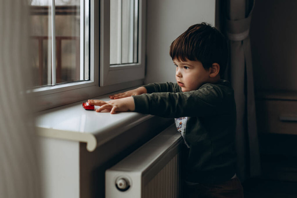 Mały, zadufany, dwuletni chłopiec siedzący przy oknie i bawiący się zabawkowym samochodem dla dzieci.  - Zdjęcie, obraz