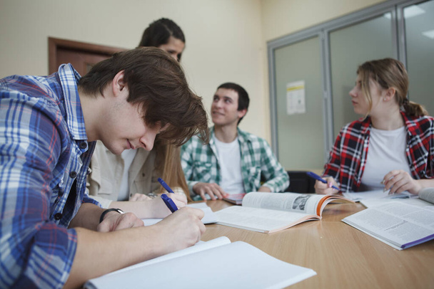Молодий чоловік пише нотатки під час занять, його однокласники говорять на фоні
 - Фото, зображення
