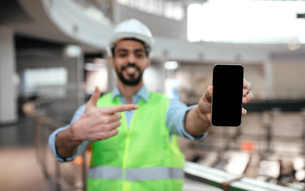 Happy millennial islamic engineer male in protective uniform, hard hat with beard point finger at phone with empty screen in factory interior. Ad, app and offer for business, industry, factory control - Photo, Image