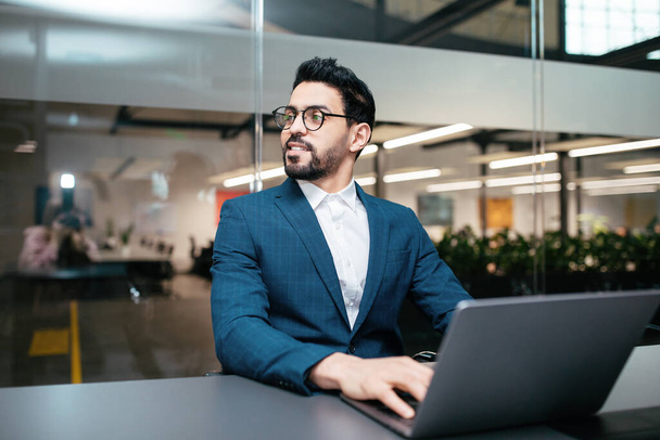 Serious busy attractive millennial muslim businessman with beard in suit, glasses works with pc in office interior and looks at empty space. Startup online, business, industry, finance during covid-19 - Photo, Image
