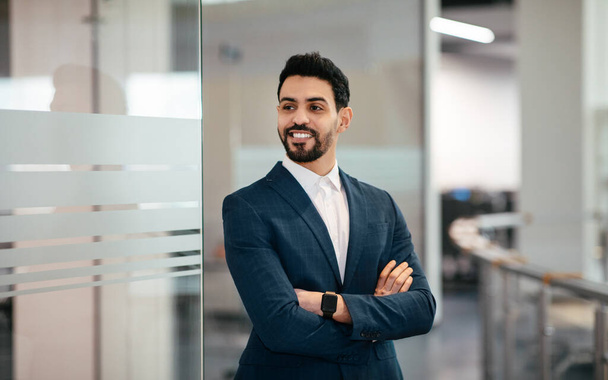 Cheerful confident handsome millennial muslim businessman with beard in suit with crossed arms looks at empty space in office interior. Business, industry, finance and startup during covid-19 outbreak - Zdjęcie, obraz