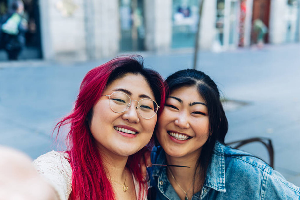 Asian young women friends smiling and having fun taking a selfie in the city. Oriental sisters taking a photo outdoors. Travel, tourism and family vacation concept. - Foto, afbeelding