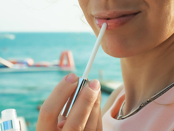 Een vrouw schildert haar lippen met lipgloss op zomerse dag tegen de achtergrond van de zee. Een close-up foto. Zachte focus. - Foto, afbeelding