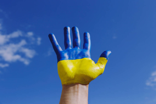 Close up shot of a man's hand lit by sunshine painted in colors of Ukrainian national flag. .A hand raised over blue sky background as a symbol of support and solidarity with Ukraine. Copy space. - Foto, imagen