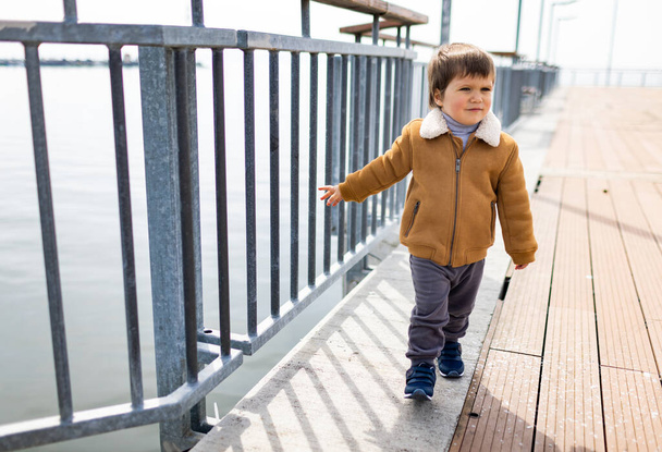 A little happy cool boy in stylish fashionable warm clothes walks along a wide empty pier with an old rusty gray fence near the calm quiet big Black Sea, in autumn cloudy weather - Zdjęcie, obraz