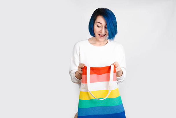 Mujer con el pelo azul sosteniendo una bolsa de arco iris sobre un fondo blanco. Bandera LGBT colores. - Foto, Imagen