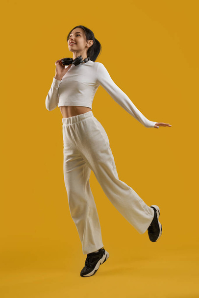 Inspired graceful lady dancing, with headphones around neck in studio. Front view of happy young girl in casual clothes, jumping with earphones, isolated on orange background. Concept of inspiration. - Fotoğraf, Görsel