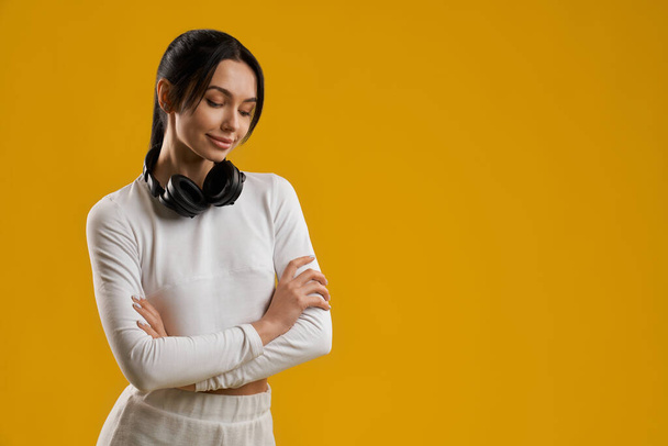 Dreamy brunette female with ponytail standing in crossed-arms pose in studio. Portrait view of smiling girl with black headphones looking down, isolated on orange background. Concept of lifestyle. - Фото, зображення