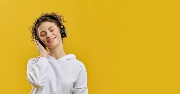 Relaxed female with black earmuffs listening music, having rest indoor. Portrait view of smiling girl pressing headphone to ear, enjoying sound, isolated on orange background. Concept of music lover. - Фото, зображення