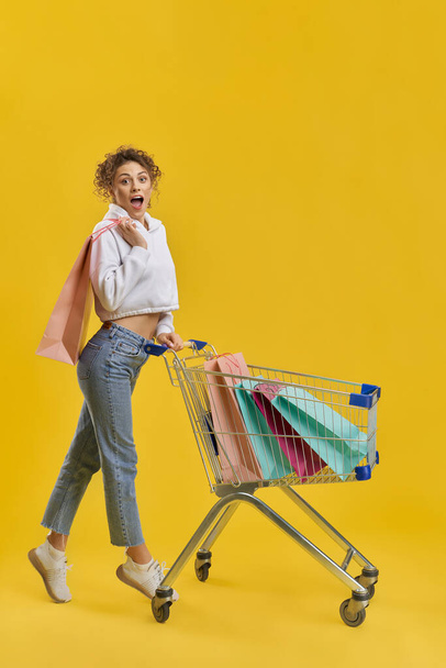 Shocked woman carrying shopping cart, with bright paper bags inside. Side view of surprised girl with shopping trolley, standing on tiptoe, isolated on orange studio background. Concept of shopping.  - Foto, Imagem