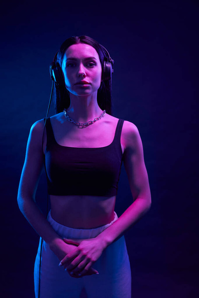 Confident woman in sporty outfit standing with folded arms in studio. Portrait view of serious girl wearing black headphones looking at camera, illuminated with magenta light. Concept of lifestyle. - Foto, Bild
