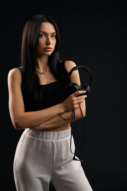 Confident lady holding headphones, while standing in studio. Front view of dreamy girl in sporty wear holding arms crossed, while looking at camera, isolated on black background. Concept of lifestyle. - Φωτογραφία, εικόνα