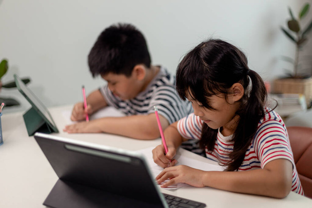 Asiatiques garçons et filles profiter de l'apprentissage en ligne en prenant des notes et en utilisant des tablettes à la maison. - Photo, image