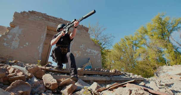 A Ukrainian soldier with a rifle pointed in front of him leaves the destroyed house and aims at the sides. - Photo, Image