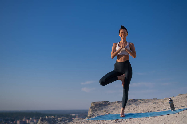 Красивая стройная кавказка в спортивной одежде практикует йогу на скале на фоне голубого неба на закате - Фото, изображение