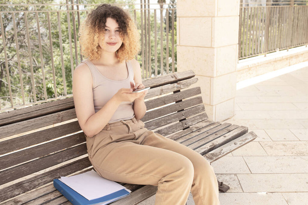 Cute curly teenage girl holds a phone in her hand, chats while sitting on bench. Schoolgirl, student writes sms online. Lifestyle. Outside. Concept of digital communications, virtual love. Young woman - Fotó, kép