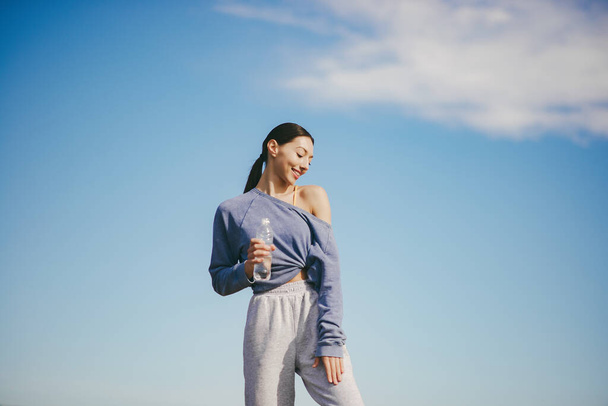 Beautiful girl training on a sky background. Woman in a blue sweater. Lady make a yoga. Girl with a bootle of water - Photo, Image