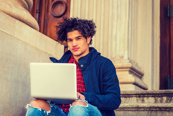 Étudiant américain avec visage roux, cheveux longs bouclés, veste bleue avec capuche, jeans détruits, assis sur les escaliers du campus à New York, travaillant sur un ordinateur portable, pensant.  - Photo, image
