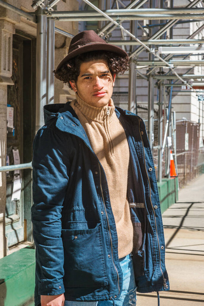 American Man traveling, walking through sidewalk bridge in New York. Wearing blue jacket with hood, knitted turtleneck sweater, Fedora hat, guy with freckle face, curly hair, looking at you under sun. - Foto, imagen