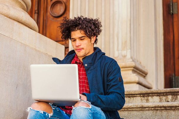 Étudiant américain avec visage roux, cheveux longs bouclés, veste bleue avec capuche, jeans détruits, assis sur les escaliers sur le campus de New York, lecture, travail sur ordinateur portable.  - Photo, image