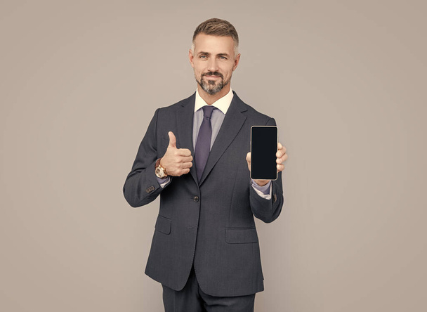 mature man influencer with grizzled hair in suit presenting mobile phone device and show thumb up, best offer. - Фото, зображення