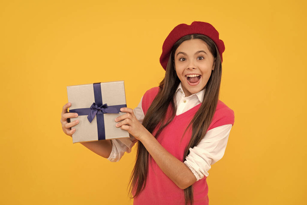 surprised teen girl in french beret hold present or gift box on yellow background, purchase. - 写真・画像