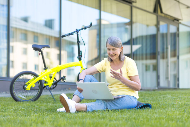 Education online. Portrait of senior beautiful woman sitting on campus on grass with laptop and bicycle. Talking on a video call on a laptop, waving his arms, smiling - Φωτογραφία, εικόνα