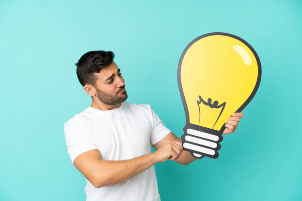 Young handsome caucasian man isolated on blue background holding a bulb icon and having doubts - Foto, immagini