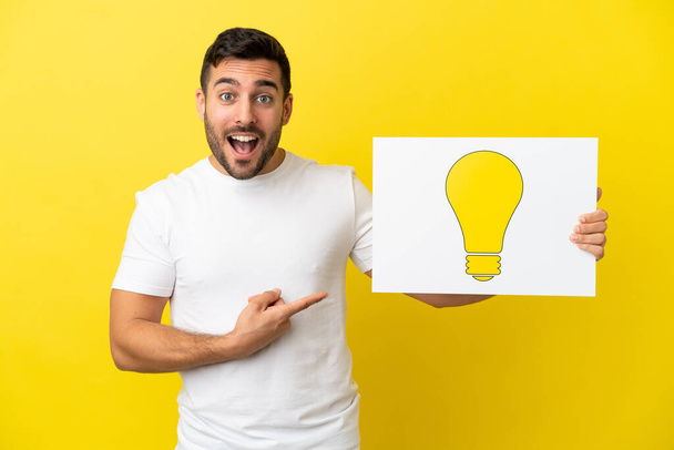 Young handsome caucasian man isolated on yellow background holding a placard with bulb icon and pointing it - Foto, Imagem
