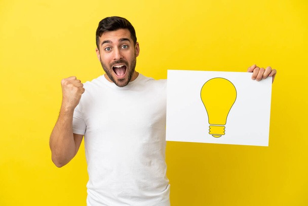 Young handsome caucasian man isolated on yellow background holding a placard with bulb icon and celebrating a victory - Foto, Imagem