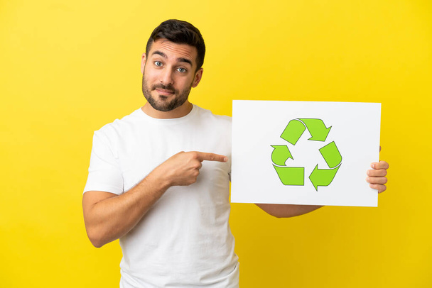 Joven hombre caucásico guapo aislado sobre fondo amarillo sosteniendo un cartel con icono de reciclaje y señalándolo - Foto, imagen