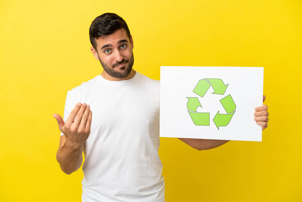 Junger gutaussehender kaukasischer Mann isoliert auf gelbem Hintergrund mit einem Plakat mit Recycling-Symbol und kommender Geste - Foto, Bild