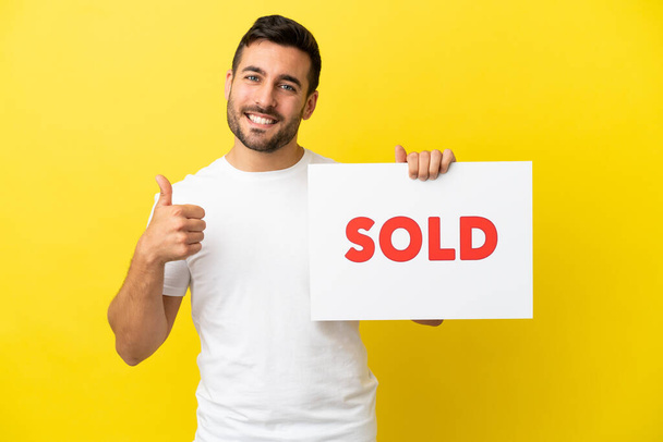 Young handsome caucasian man isolated on yellow background holding a placard with text SOLD with thumb up - Fotografie, Obrázek