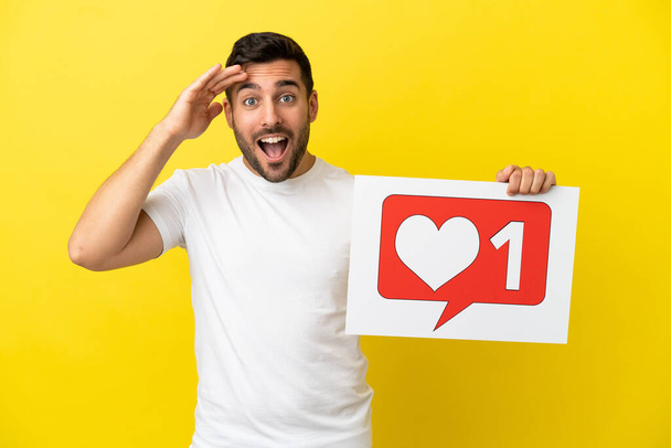 Young handsome caucasian man isolated on yellow background holding a placard with Like icon with surprised expression - Photo, image