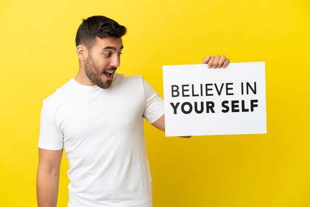 jovem bonito caucasiano homem isolado no amarelo fundo segurando um cartaz com texto Acredite em seu auto - Foto, Imagem