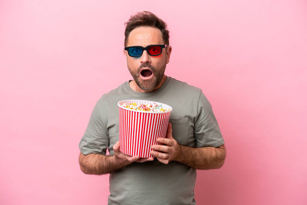 Middle age caucasian man isolated on pink background surprised with 3d glasses and holding a big bucket of popcorns - Fotoğraf, Görsel